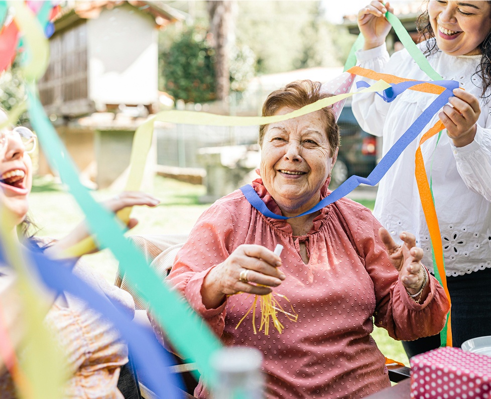 Older woman laughing among colourful streamers at an outdoor family celebration
